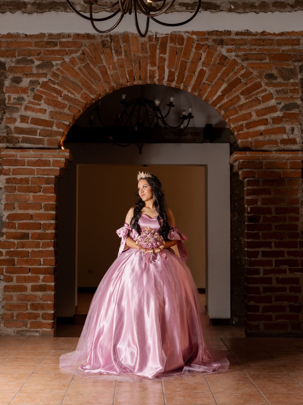 Quinceañera con vestido rosa y ramo de flores bajo un arco de ladrillo